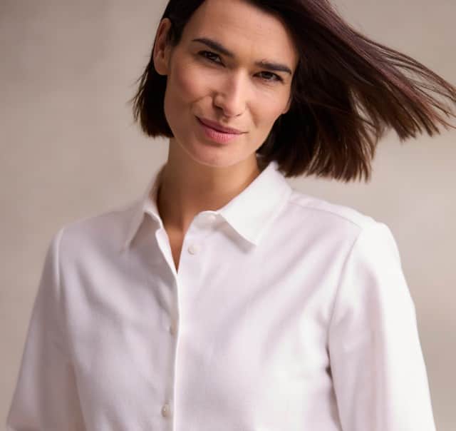 Eine Frau mit schulterlangem, braunem Haar trägt eine weiße Bluse und blickt mit leichtem Lächeln und wehendem Haar in die Kamera.