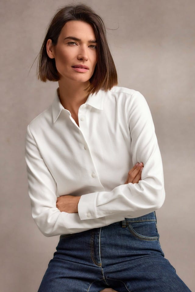 Eine Frau mit schulterlangem, braunem Haar trägt eine weiße, langärmlige Bluse und eine dunkelblaue Jeans, sitzt leicht nach vorne gelehnt und blickt ruhig in die Kamera.
