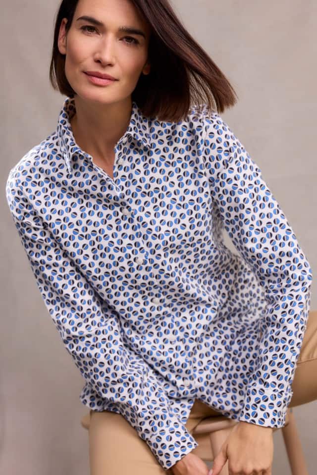 Eine Frau mit schulterlangem, braunem Haar trägt eine weiße Bluse mit blauem Muster und eine beige Hose, sitzt leicht nach vorn gebeugt und blickt ruhig in die Kamera.