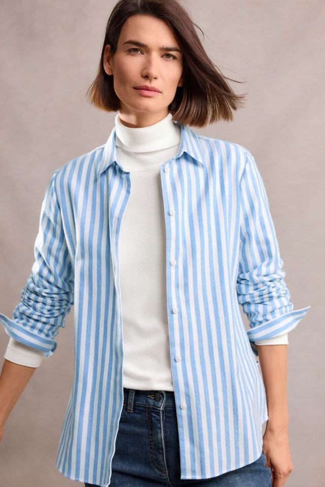 Eine Frau mit schulterlangem, braunem Haar trägt ein weißes Rollkragenshirt, darüber eine hellblau-weiß gestreifte Bluse und eine dunkelblaue Jeans, und blickt ruhig in die Kamera.