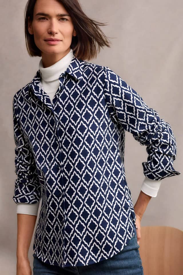 Eine Frau mit schulterlangem, braunem Haar trägt ein weißes Rollkragenshirt unter einer dunkelblauen Bluse mit weißem Rautenmuster und blickt ruhig in die Kamera.