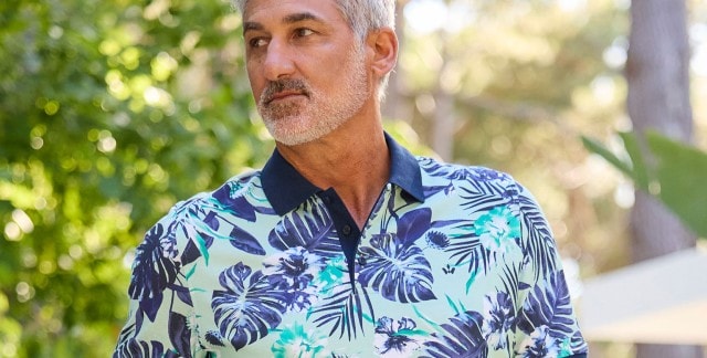 Ein Mann trägt ein kurzärmliges, gemustertes Polo-Shirt mit tropischen Blumen und Blättern.
