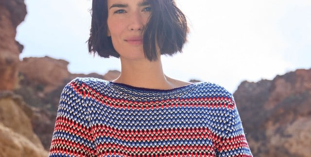 Eine Frau steht vor Felsen und trägt einen Baumwoll-Pullover Nordic Stripe.