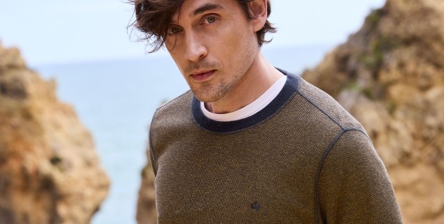 Ein Mann steht an einer Felsküste und trägt einen Struktur Pullover. 