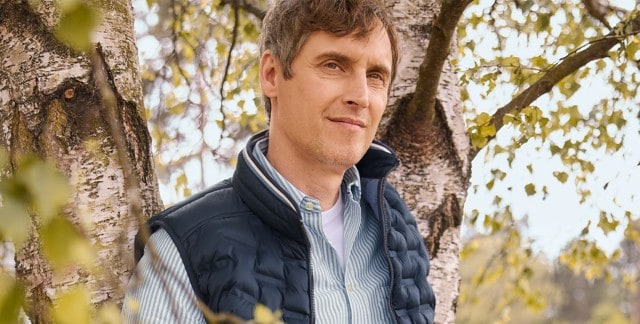 Ein Mann lehnt an einem Baum und trägt eine dunkelblaue Steppweste über einem hell gestreiften Hemd mit weißem T-Shirt darunter.