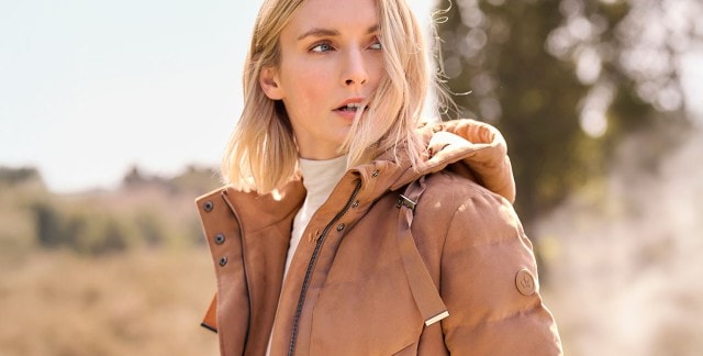 Eine Frau trägt eine hellbraune, wattierte Jacke mit Kapuze, Reißverschluss und breiten Bändern im modernen, schlichten Stil über einem hellen Rollkragenpullover. Eine Frau trägt eine hellbraune, wattierte Jacke mit Kapuze, Reißverschluss und breiten Bändern im modernen, schlichten Stil über einem hellen Rollkragenpullover.