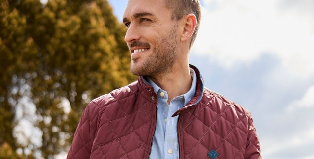 Eine Person trägt eine gesteppte, weinrote Jacke über einem hellblauen Hemd und steht im Freien vor einem unscharfen, grünen Hintergrund.