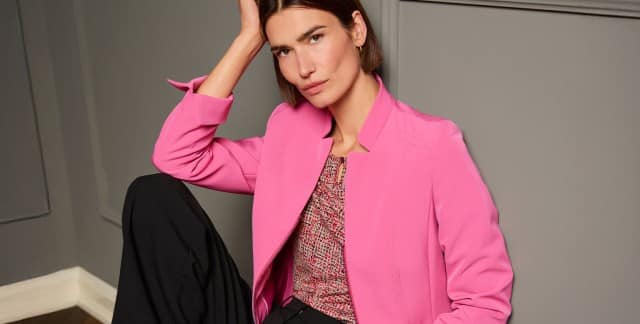 Eine Frau sitzt lässig auf einem Stuhl und trägt den Kofferblazer Airline in Soft Pink, darunter ein Viskoseshirt in Bouclelook und dazu eine schwarze Hose. Eine Frau sitzt lässig auf einem Stuhl und trägt den Kofferblazer Airline in Soft Pink, darunter ein Viskoseshirt in Bouclelook und dazu eine schwarze Hose.