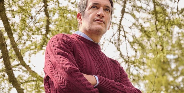 Ein Mann steht vor einem Baum und trägt den Aran-Pullover in Bordeaux, darunter ein hellblaues Hemd.
