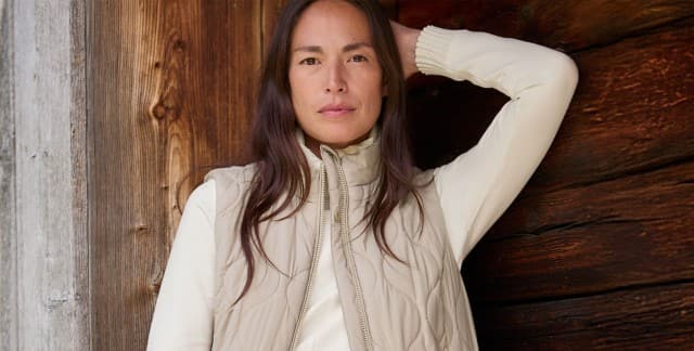 Eine Frau steht vor einer Holzwand und trägt die Wellensteppweste in Toffee/Creme, darunter ein weißes Sweatshirt und dazu eine Jeans.