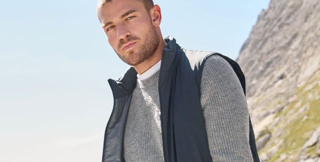 Ein Mann gesteht vor einem Gebirge und trägt eine Weste in Marine, darunter einen grauen Pullover. Ein Mann gesteht vor einem Gebirge und trägt eine Weste in Marine, darunter einen grauen Pullover.