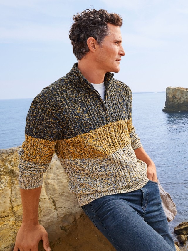 Ein Mann trägt einen gestrickten Troyer und steht an den Felsen einer Küste.