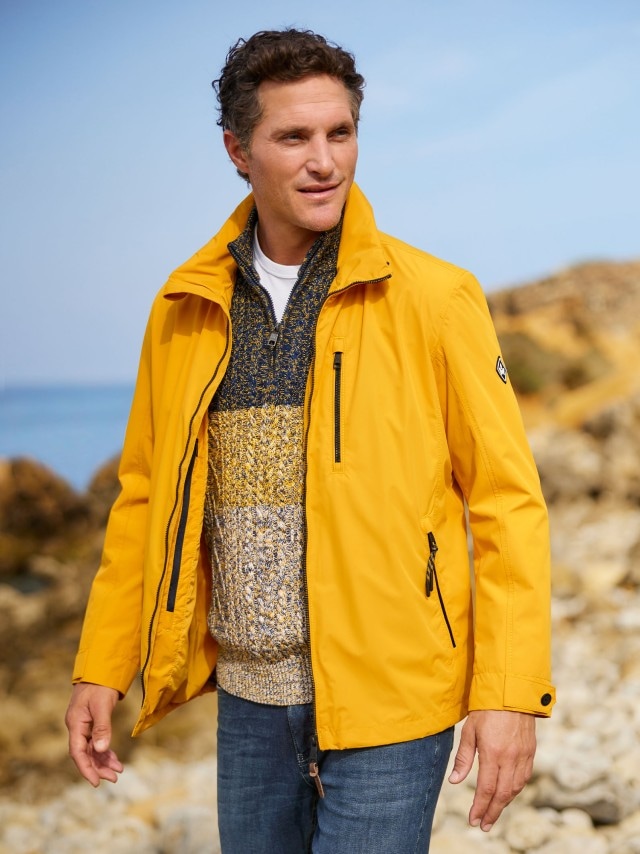 Ein Mann trägt eine gelbe Wetterschutzjacke und steht an den Felsen einer Küste