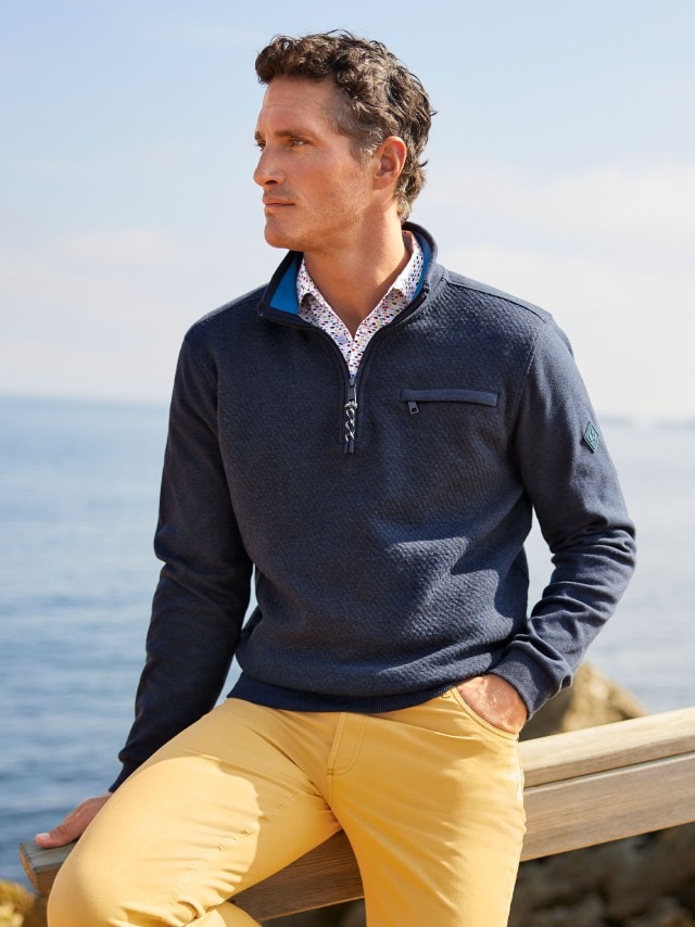 Ein Mann trägt einen blauen Troyer und eine gelbe Hose und steht an einem Steg am Strand.