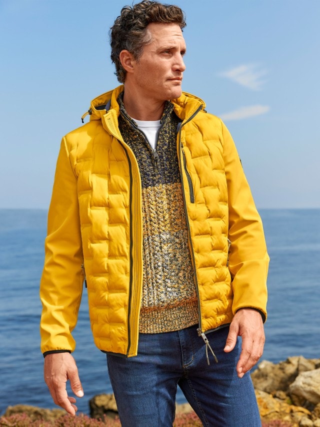 Ein Mann steht im gelben Blouson und Strick-Troyer auf den Felsen einer Küste