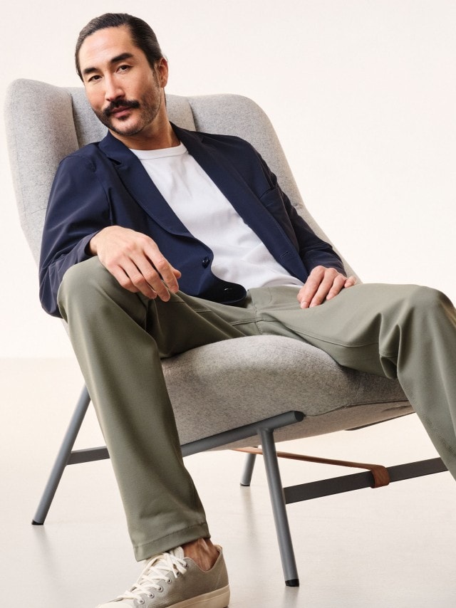 Ein Mann sitzt auf einem Sessel und trägt ein dunkelblaues Sakko, grüne Hose und weißes Shirt.