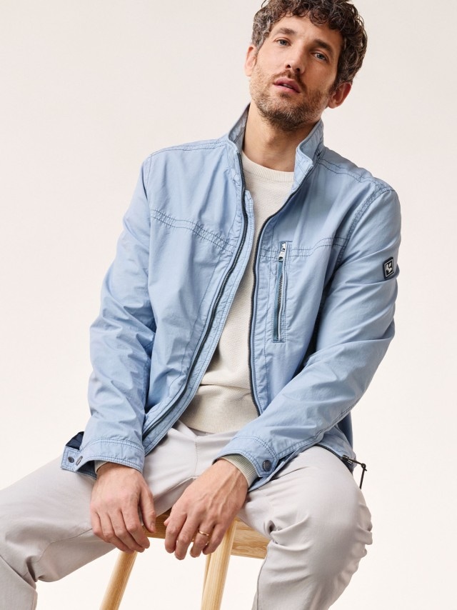 Ein Mann sitzt auf einem Stuhl und trägt einen hellblauen Blouson und eine Chino in Safaribeige