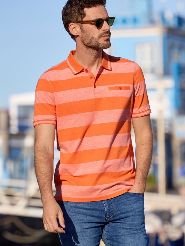 Ein dunkelhaariger Mann mit Sonnenbrille in einem Polo-Shirt mit orangefarbenen Streifen.