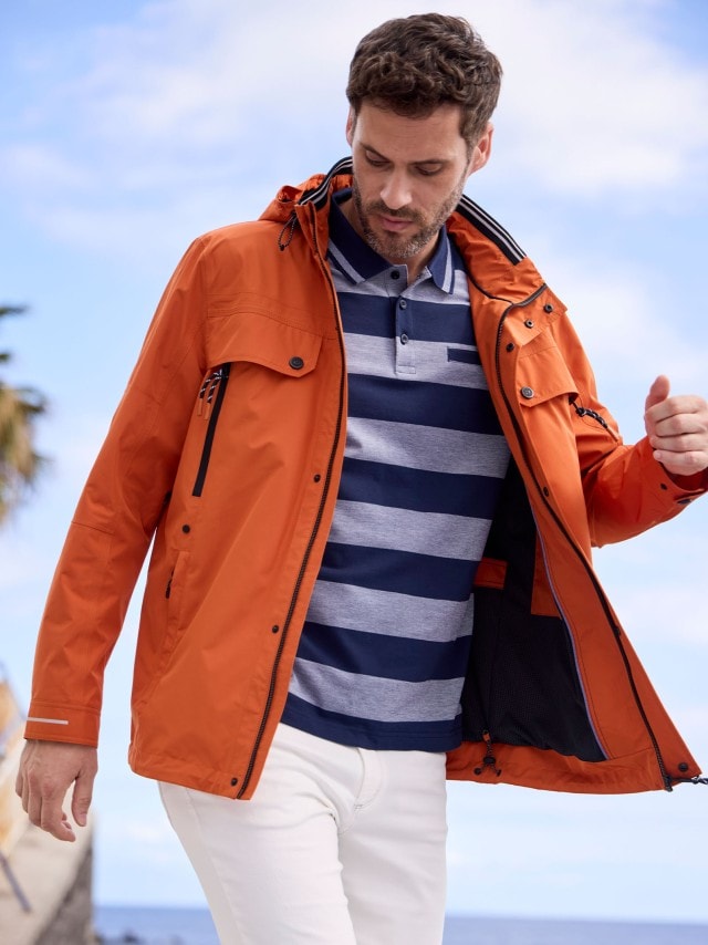 Dunkelhaariger Mann in schwungvoller Pose trägt ein gestreiftes Polo-Shirt, eine weiße Hose und eine Aquastop Reisejacke in Orange.