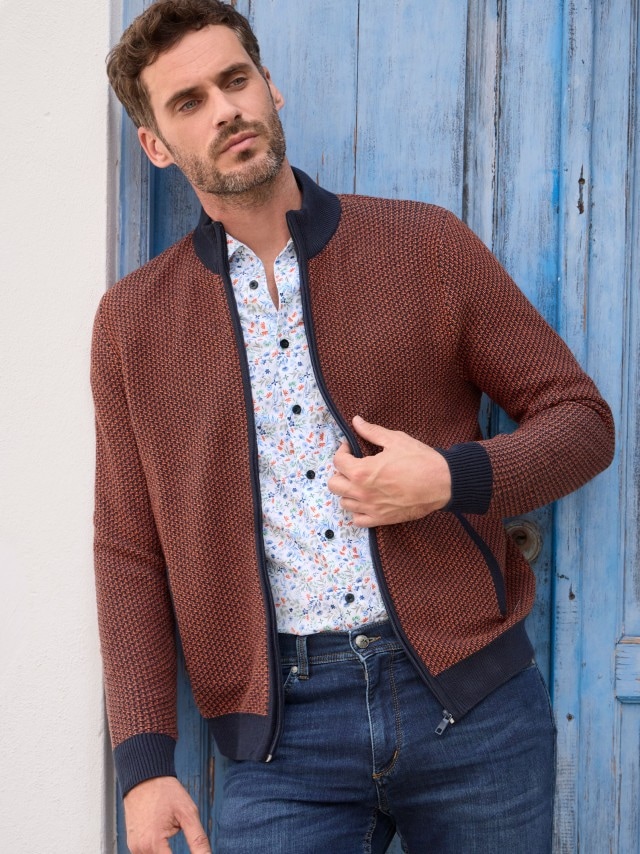 Ein Mann in Zip-Strickjacke, geblümtem Hemd und Jeans steht vor einer blauen Holzwand