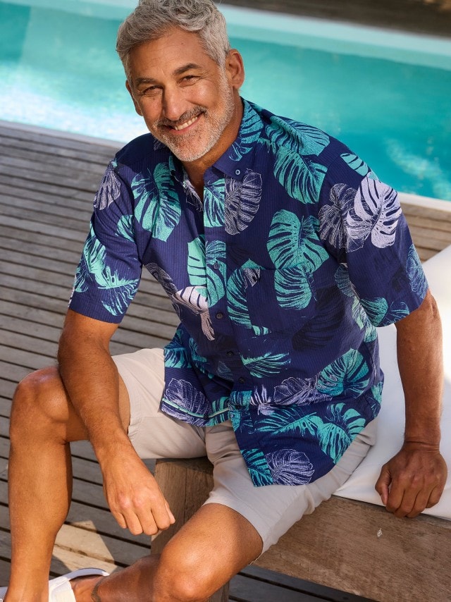 Ein Mann sitzt entspannt am Rand eines Swimmingpools und trägt ein kurzärmliges, blaues Hemd mit grünem und weißem Blattmuster sowie beige Shorts.