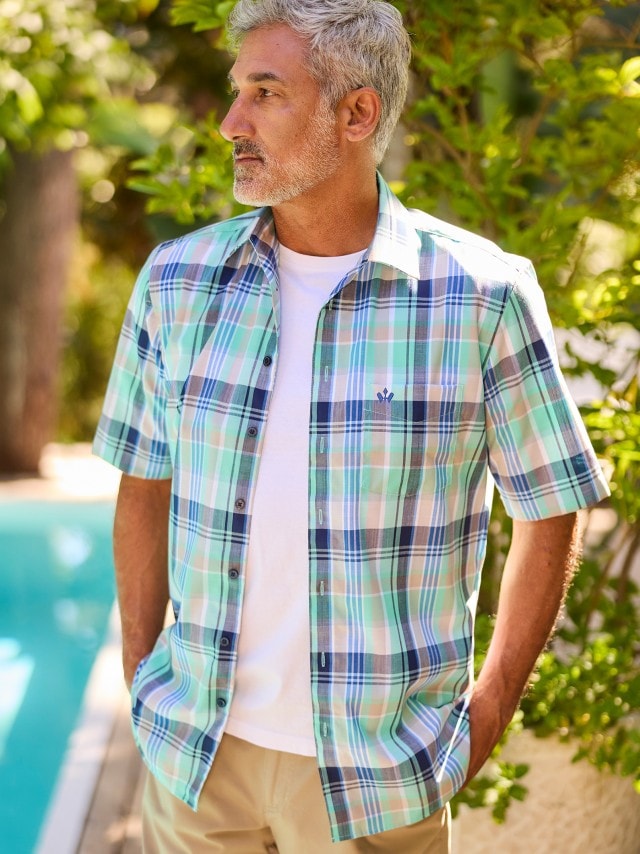 Ein Mann steht im Freien und trägt ein kurzärmeliges Hemd mit grün-blauem Karomuster über einem weißen T-Shirt sowie eine beige Hose.