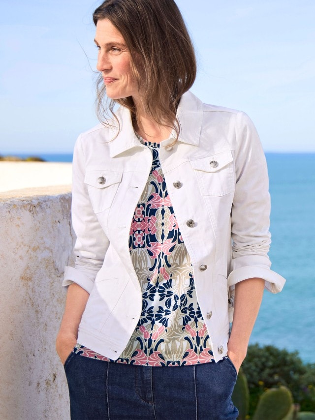 Eine Frau trägt eine weiße Jeansjacke über einem gemusterten Shirt und eine dunkelblaue Hose. Das Outfit wirkt modern und lässig.