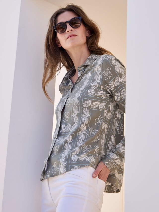 Eine Frau mit Sonnenbrille, die eine grau-grüne Bluse mit weißem floralem Muster trägt, dazu eine weiße Hose.