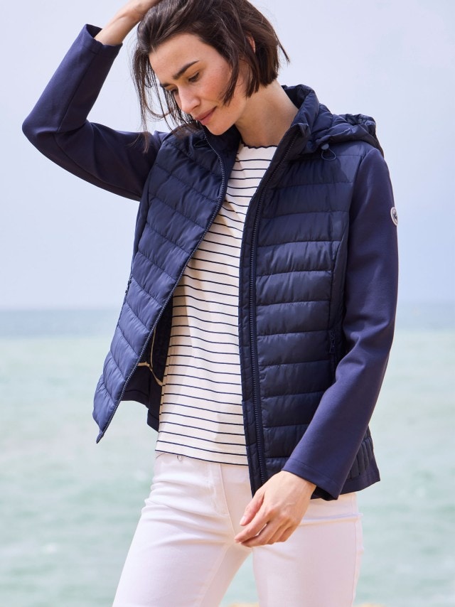 Eine Frau, die eine dunkelblaue Steppjacke, ein weißes Shirt mit dunklen Streifen und eine weiße Hose trägt.