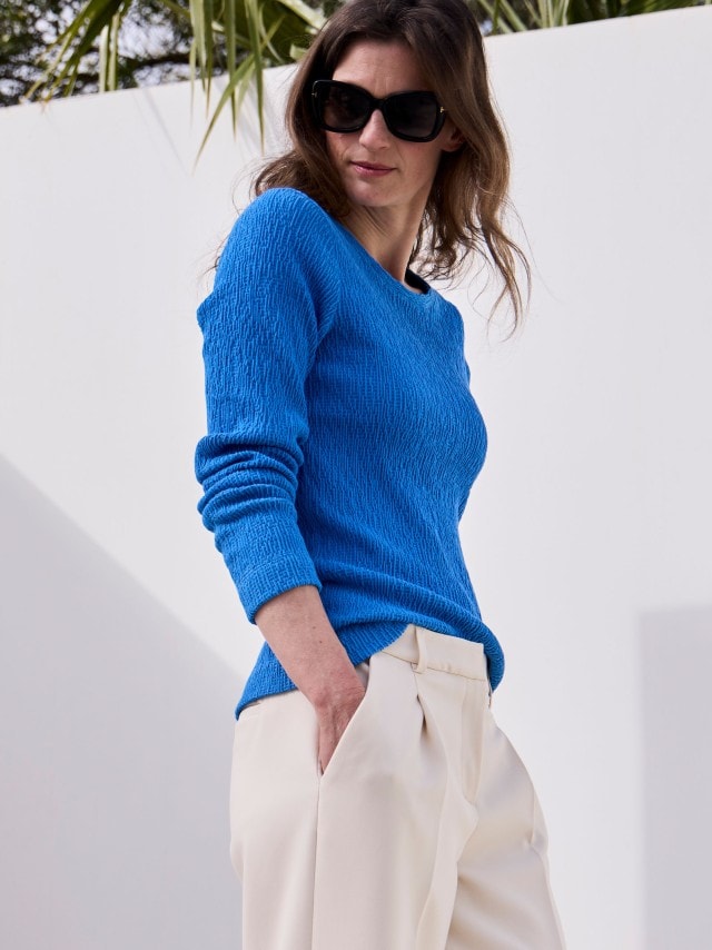 Eine Frau, die ein leuchtend blaues, strukturiertes Langarmshirt und eine weite, cremefarbene Hose trägt.