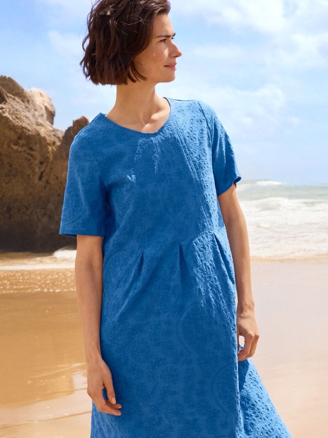 Eine Frau steht am Strand und trägt ein leuchtend blaues, kurzärmeliges Kleid mit dezentem Muster.
