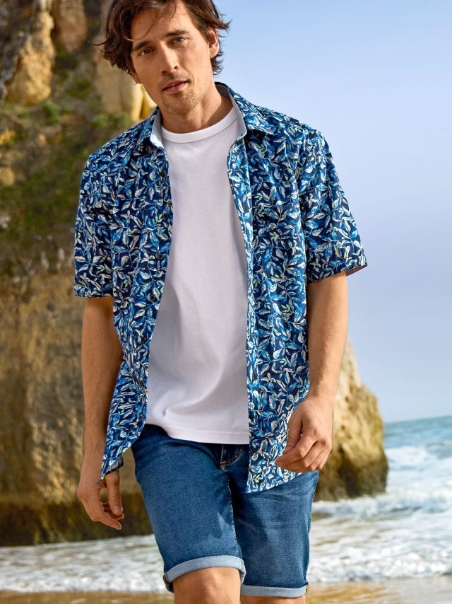 Ein Mann trägt ein weißes T-Shirt, eine blaue gemusterte Kurzarmhemd und eine blaue Jeansshorts. Der Stil ist sommerlich und entspannt, perfekt für einen Strandspaziergang.