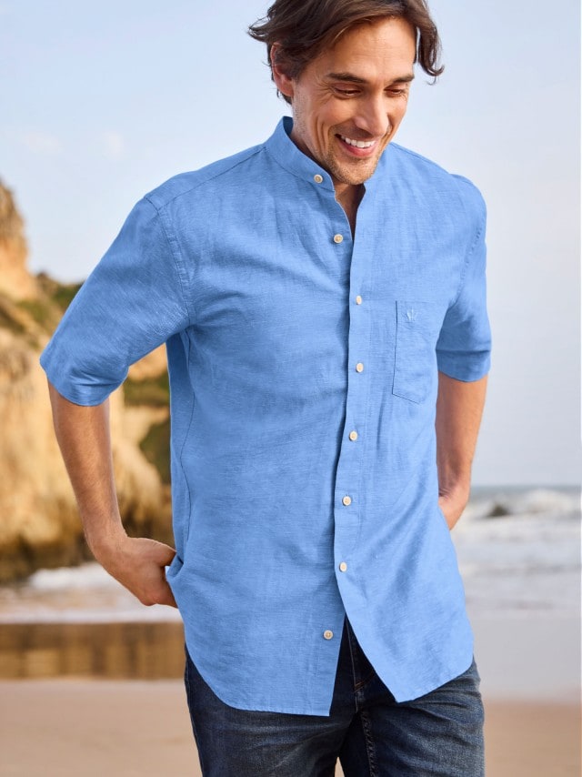 Ein Mann trägt ein hellblaues Leinenhemd mit Stehkragen und eine dunkle Jeans. Sein Look ist sommerlich-elegant und eignet sich gut für einen Tag am Strand.