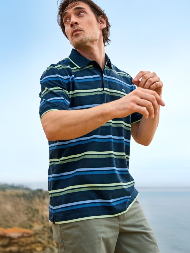 Ein Mann trägt ein gestreiftes Poloshirt in Blau- und Grüntönen sowie eine helle Hose. Der Stil ist sportlich-lässig und passt gut zu einem Tag am Meer.