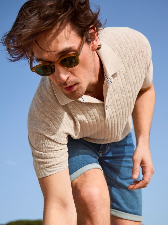 Ein Mann trägt ein beiges, geripptes Poloshirt und eine blaue Jeansshorts. Ergänzt wird der sommerlich-legere Stil durch eine Sonnenbrille.