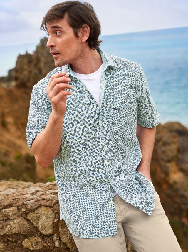 Ein Mann trägt ein gemustertes, kurzärmliges Hemd über einem weißen T-Shirt und beige Chinohosen. Der Look ist leger und sommerlich.