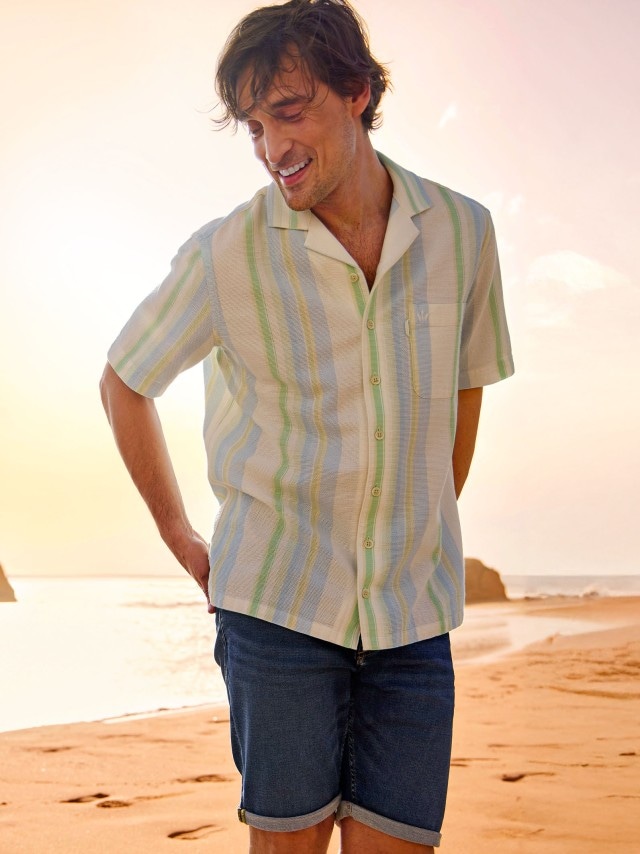 Ein Mann trägt ein kurzärmeliges, gestreiftes Hemd in Pastellfarben und eine blaue Jeansshorts. Sein Outfit wirkt sommerlich und entspannt, ideal für einen Strandspaziergang bei Sonnenuntergang.