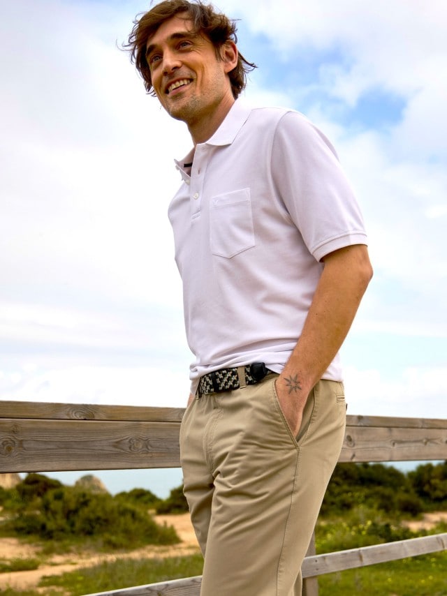 Ein Mann trägt ein weißes Poloshirt und eine beige Stoffhose mit gemustertem Gürtel. Sein Outfit wirkt klassisch und gepflegt für einen entspannten Tag im Freien.