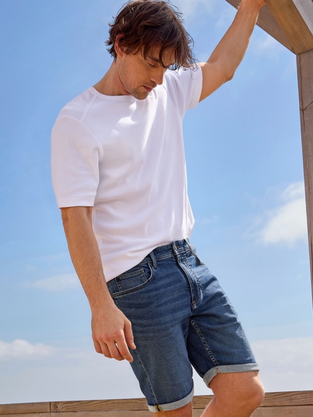 Ein Mann trägt ein weißes T-Shirt und blaue Jeansshorts mit umgekrempelten Hosenbeinen. Sein Look ist lässig und sommerlich.