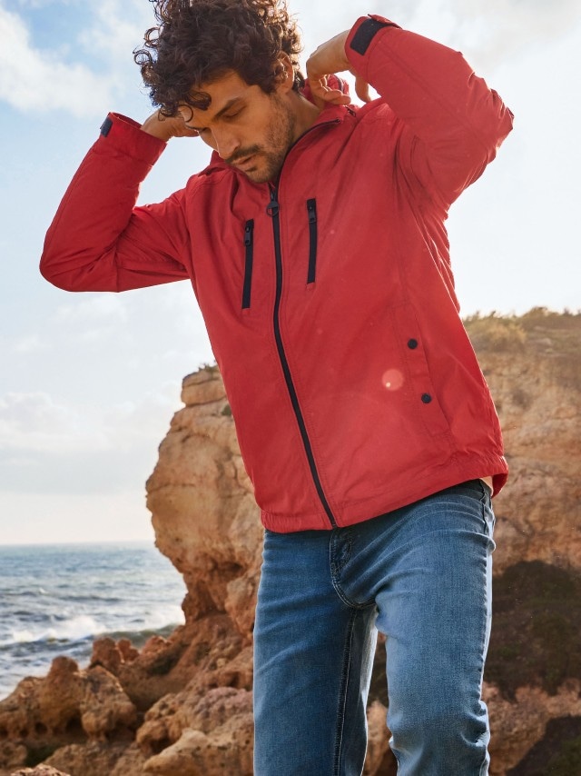 Ein Mann trägt eine rote Outdoorjacke mit schwarzen Reißverschlüssen und eine blaue Jeans. Sein Stil wirkt sportlich und lässig, passend für Aktivitäten an der Küste.