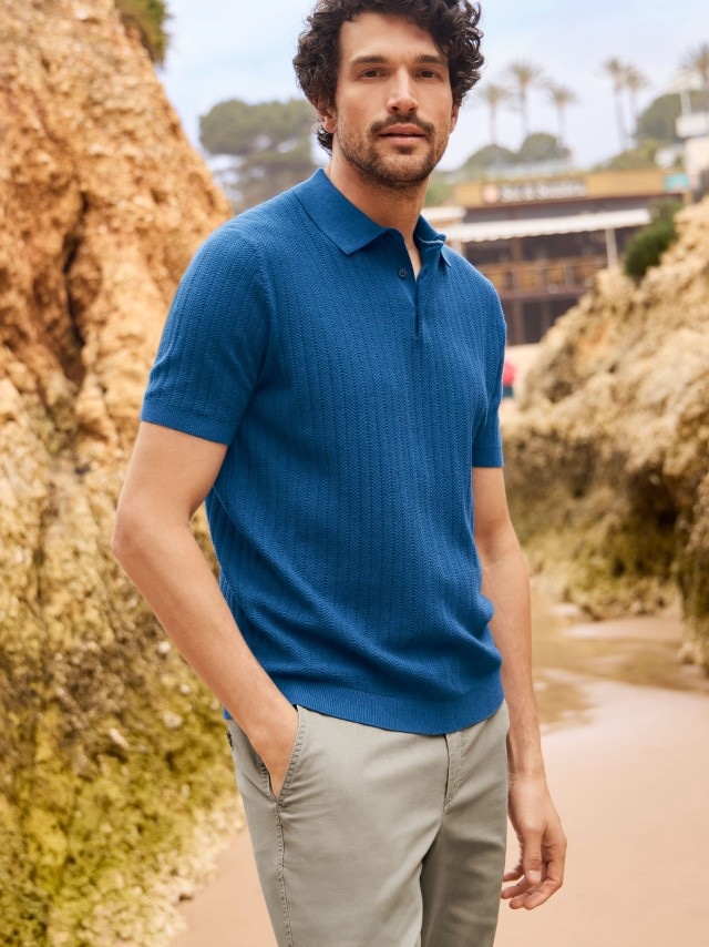 Ein Mann trägt ein blaues Strick-Poloshirt und eine beige Hose. Das Outfit sieht modern und entspannt aus.