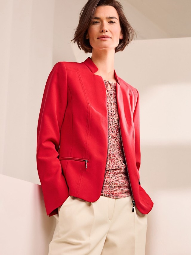 Eine Frau trägt einen roten Kofferblazer, darunter ein gemustertes rotes Shirt und dazu eine Hose in Beige.