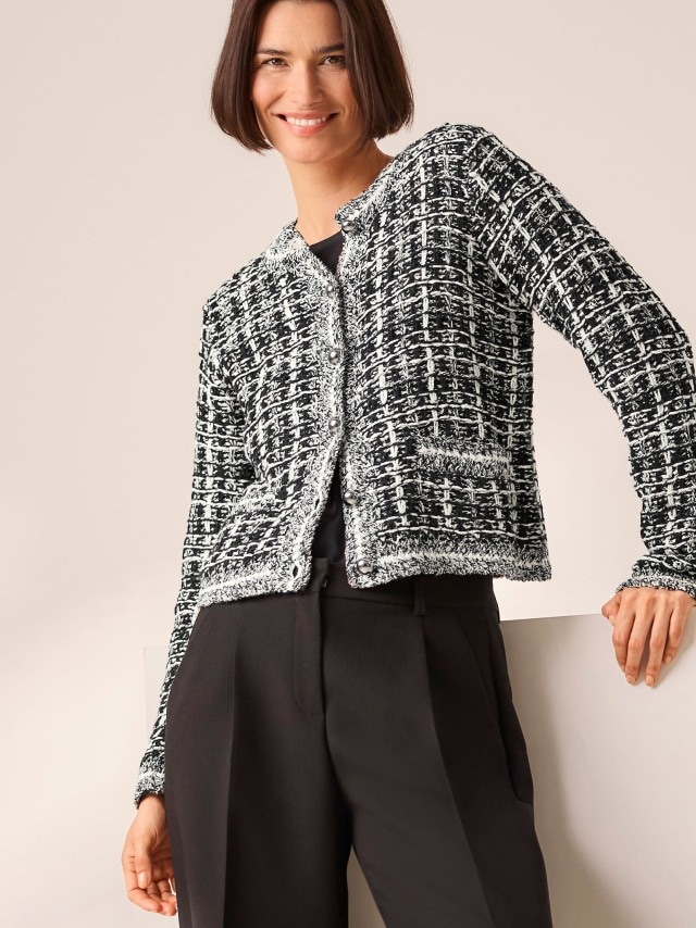 Eine Frau trägt eine schwarz-weiße Tweed Strickjacke dazu eine schwarze Hose.
