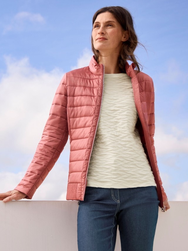 Eine Frau trägt eine Leichtsteppjacke in Rosa, darunter einen hellen Pullover und dazu eine Jeans.