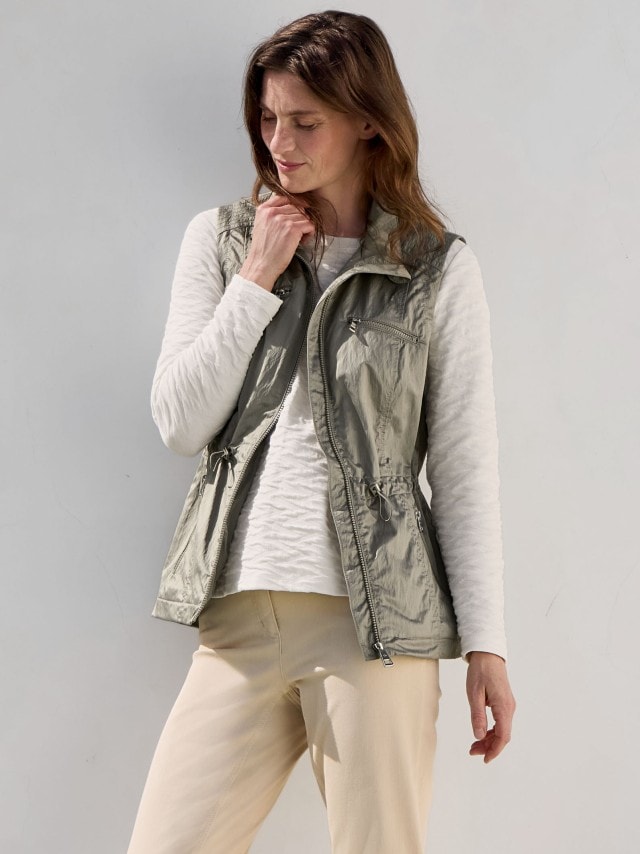 Eine Frag trägt eine Weste in Khaki, darunter ein weißes Sweatshirt und dazu eine Hose in Beige.