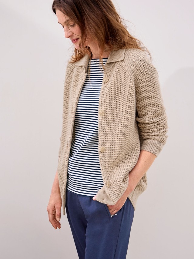 Eine Frau trägt eine Merino-Strickjacke mit Waffelstruktur in beige, darunter ein gestreiftes Shirt und dazu eine Jeans.