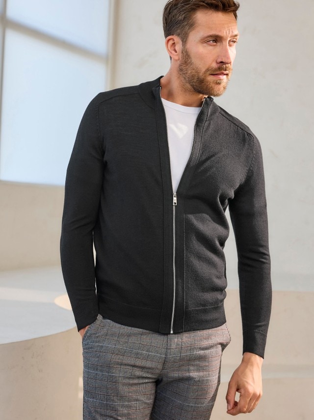 Ein Mann trägt eine Strickjacke in schwarz, darunter ein weißes Shirt und dazu eine karierte Chino.