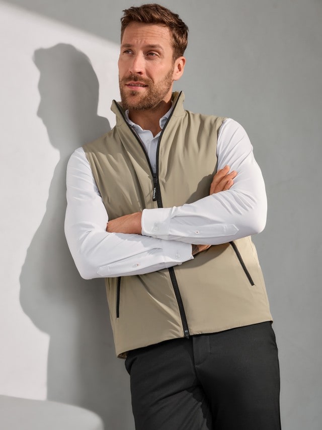 Eine Person steht mit verschränkten Armen und trägt eine beige Weste mit schwarzen Reißverschlüssen über einem weißen Hemd und einer schwarzen Hose.