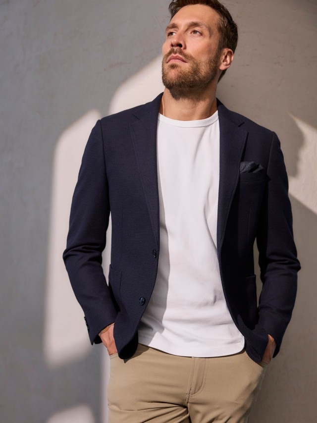 Ein Mann trägt ein Sakko in Marine, darunter ein weißes Shirt und dazu eine Hose in Beige.