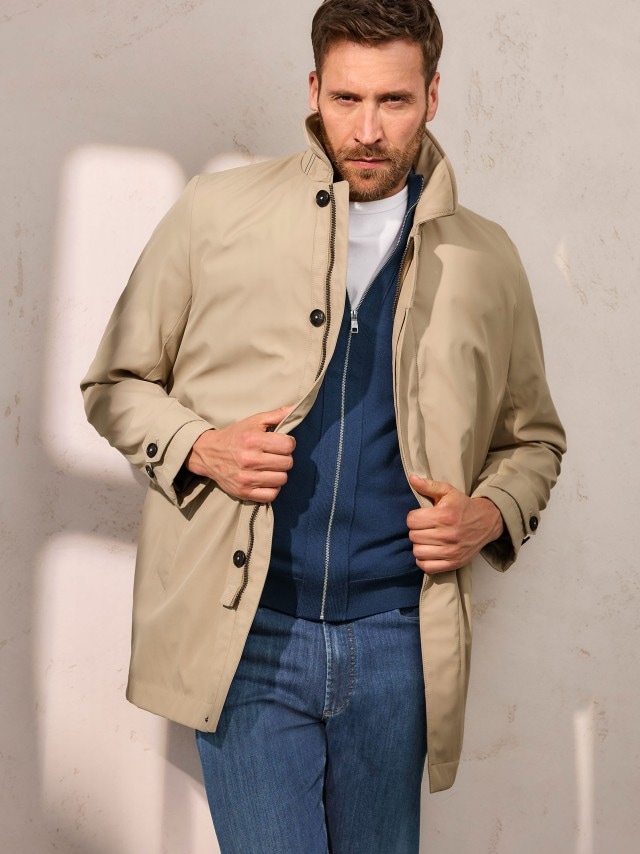 Ein Mann trägt einen Mantel in Beige, dazu eine Strickjacke sowie eine Jeans.